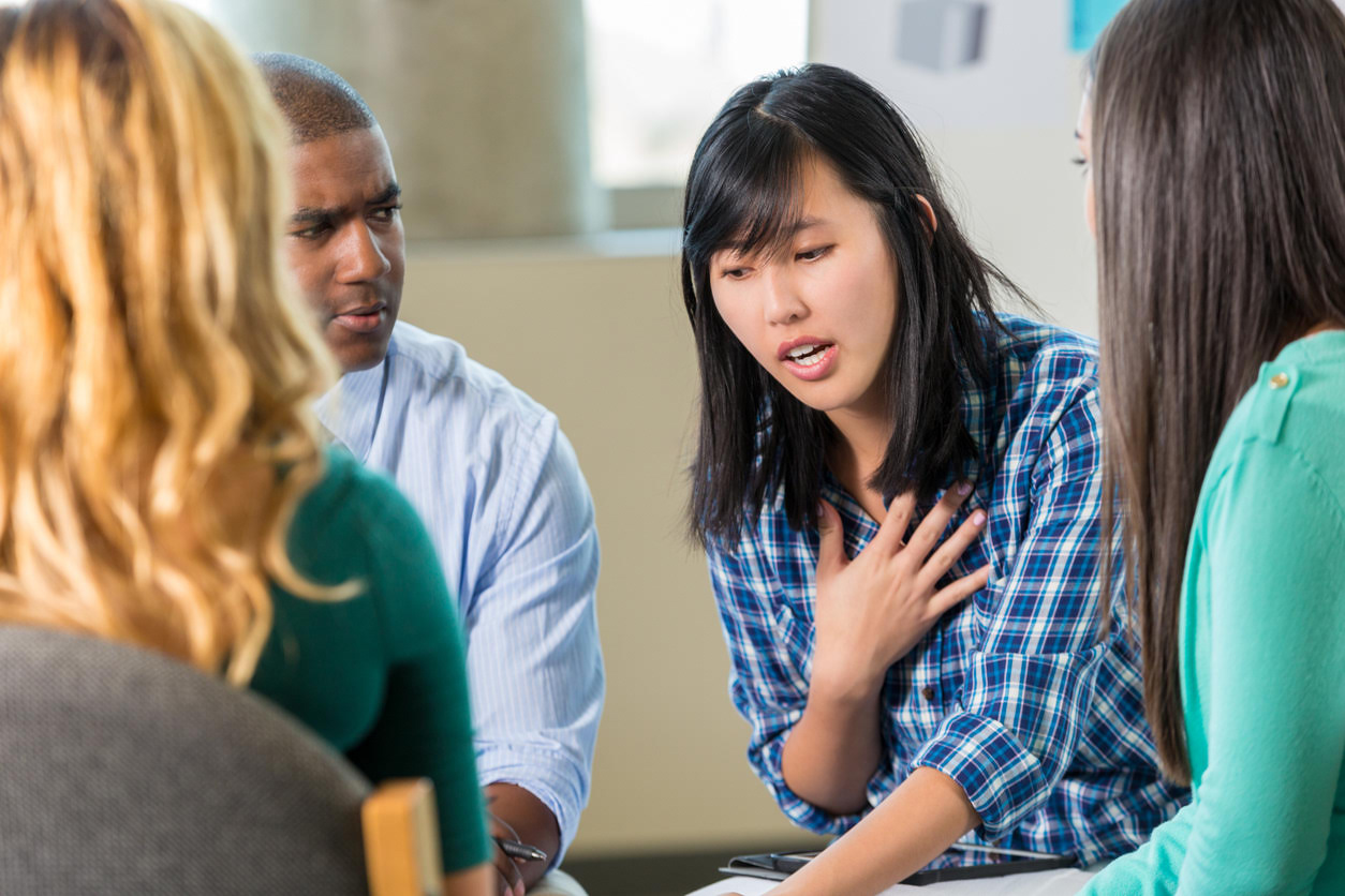 Pretty young Asian woman expresses her feelings and emotions to counselor and and support group members in therapy session. She has her hand on her chest as she is speaking. Other members are attentive and supportive toward her.